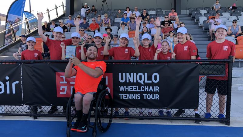 male wheelchair tennis player Dylan Alcott holds up a glass trophy in front of a group of schoolchildren