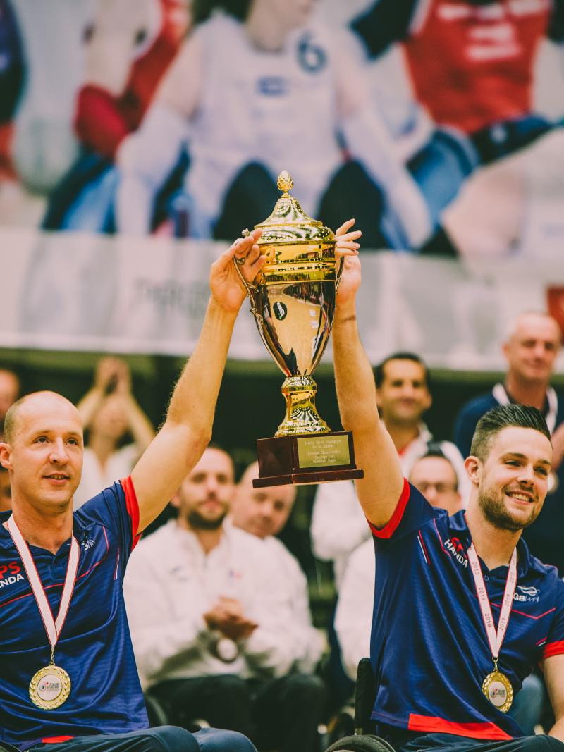 two male wheelchair rugby players with medals around their neck lift a gold cup into the air