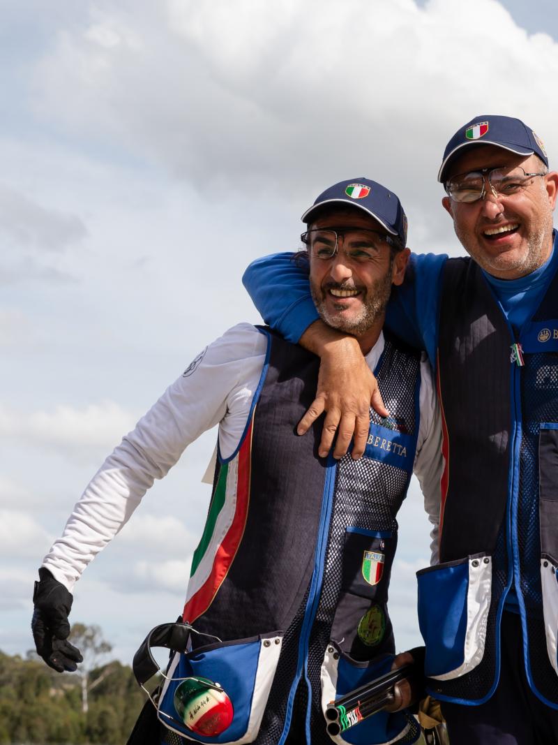 Two Italian shotgun athletes have arms around each other laughing 