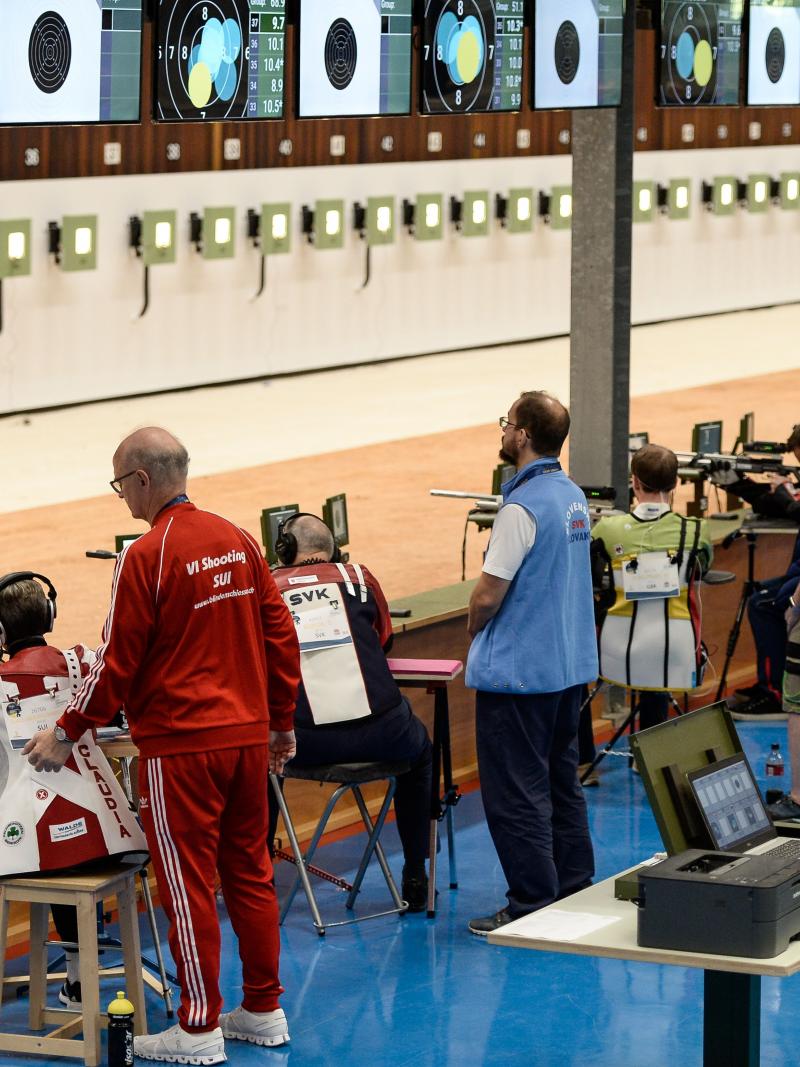 An indoor shooting range with five shooters with one person standing behind each of them