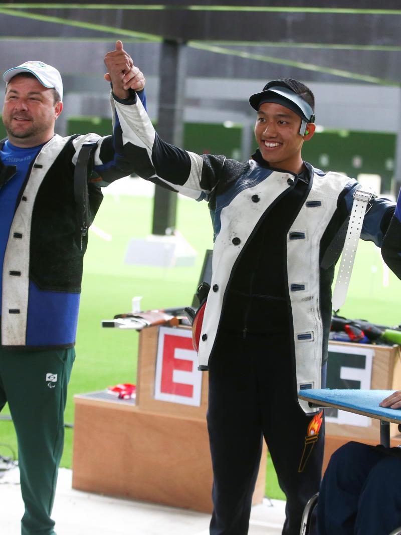 Two men standing and holding hands with a third man in a wheelchair in a shooting range