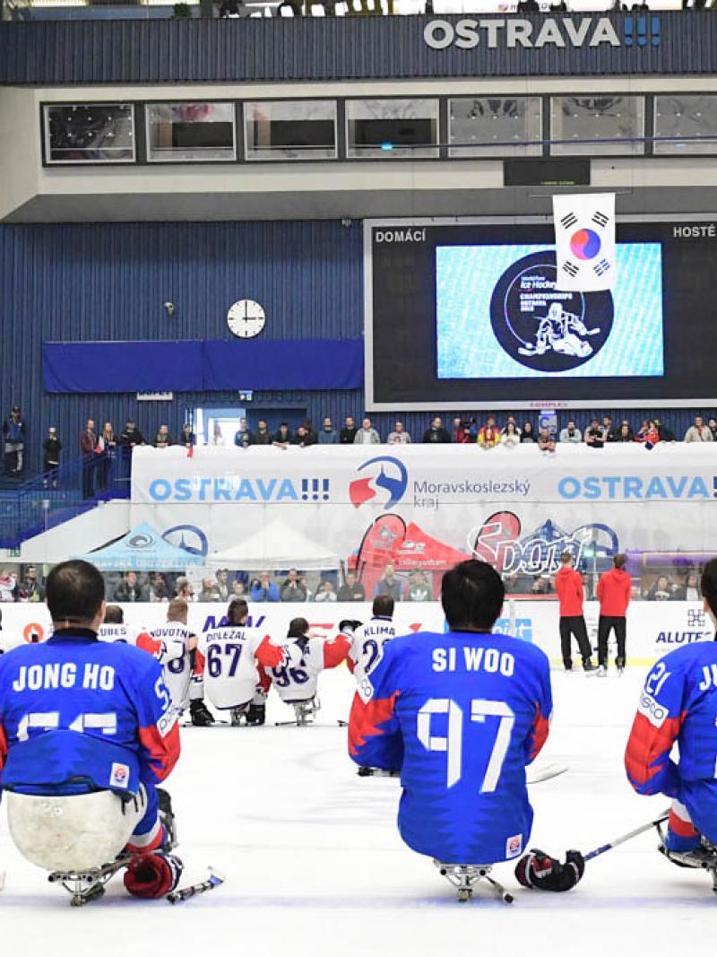 Two Para ice hockey teams on ice looking at a giant screen in a packed arena