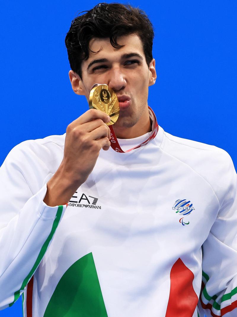 A man kisses the gold medal with a smile on his face. He is holding the miniature mascot in the other hand