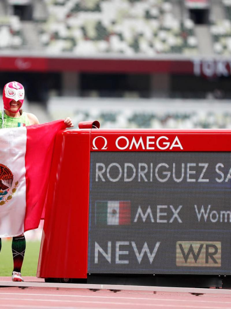 A man and a woman with Lucha Libre masks in an athletics track with the Mexican flag