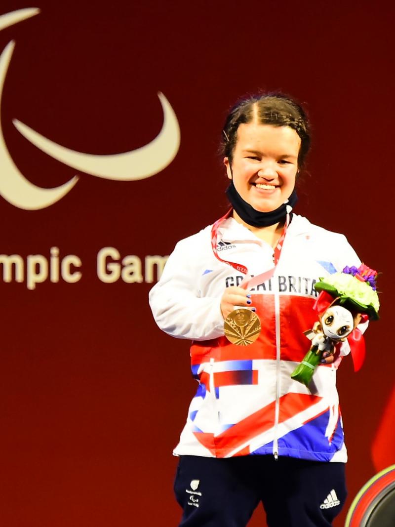 A woman holding a bronze medal in her right hand and posing for a photo on the podium.