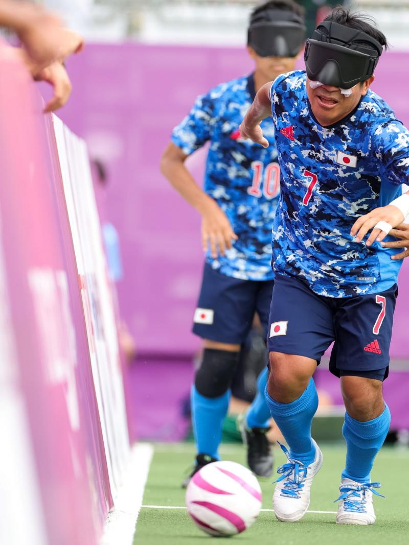 A blind football player in a blue jersey competes for the ball against a player in a white jersey at Tokyo 2020 Paralympic Games.