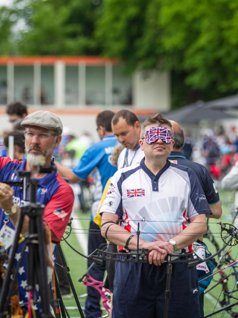 An archer in a blindfold decorated with the Union Jack rests his hands on a downturned bow.