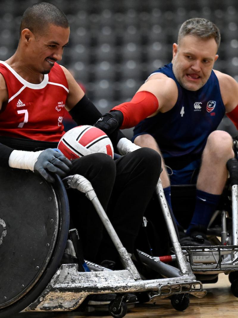 A male wheelchair rugby player wearing a red jersey carries the ball, while another player reaches for the ball.