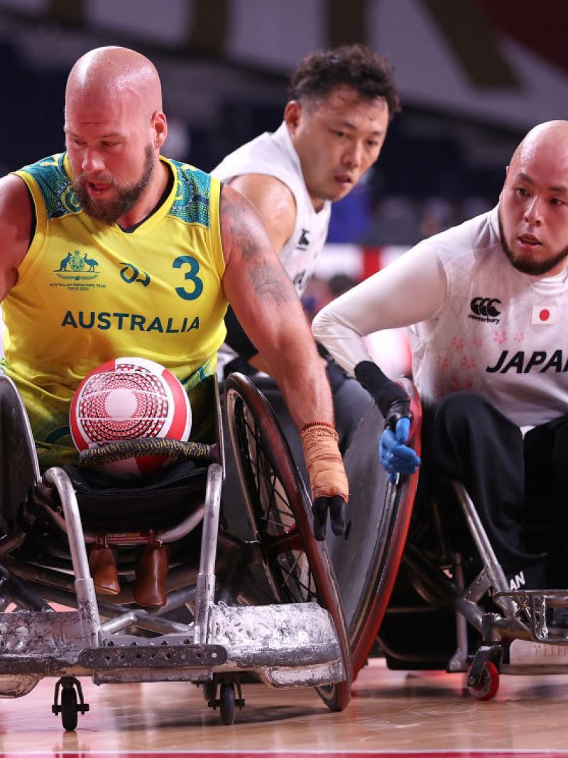 A male wheelchair rugby player in a yellow jersey carries the ball, chased by two players.