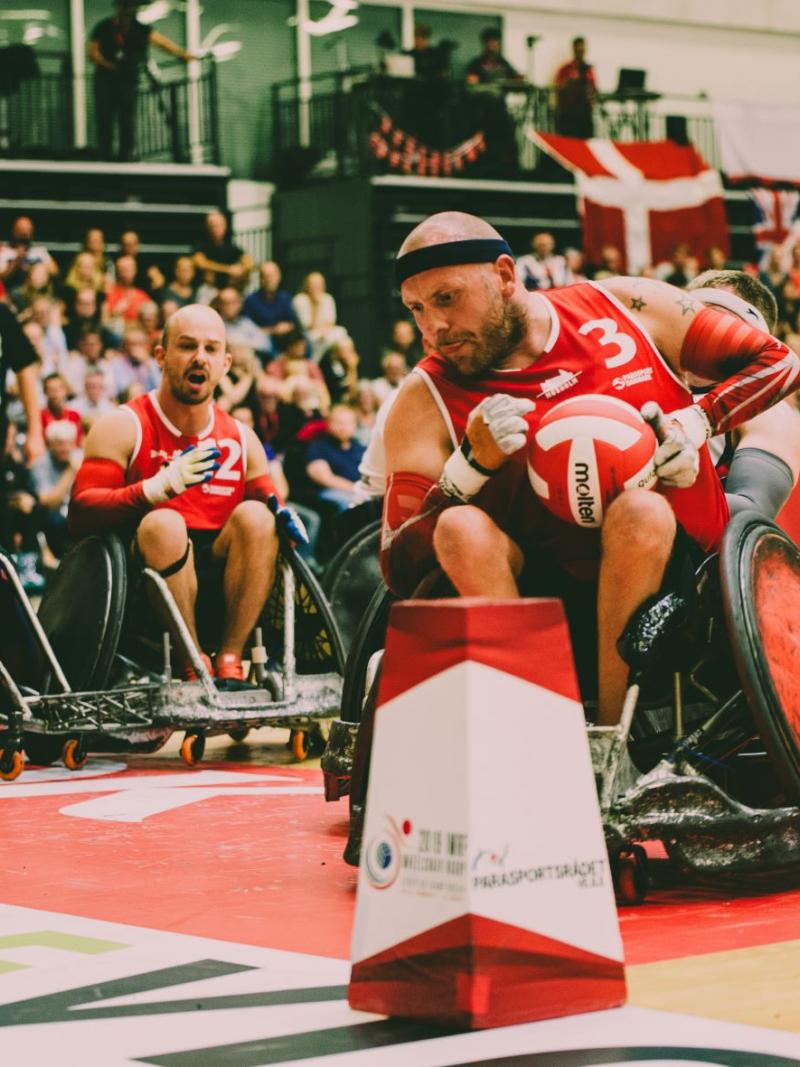 Three male athletes in wheelchairs, with one carrying a red and white ball, play at a venue packed with spectators.