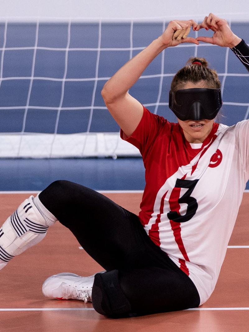 A female goalball player in an eye mask makes a heart gesture with her hands as she sits on the competition field.