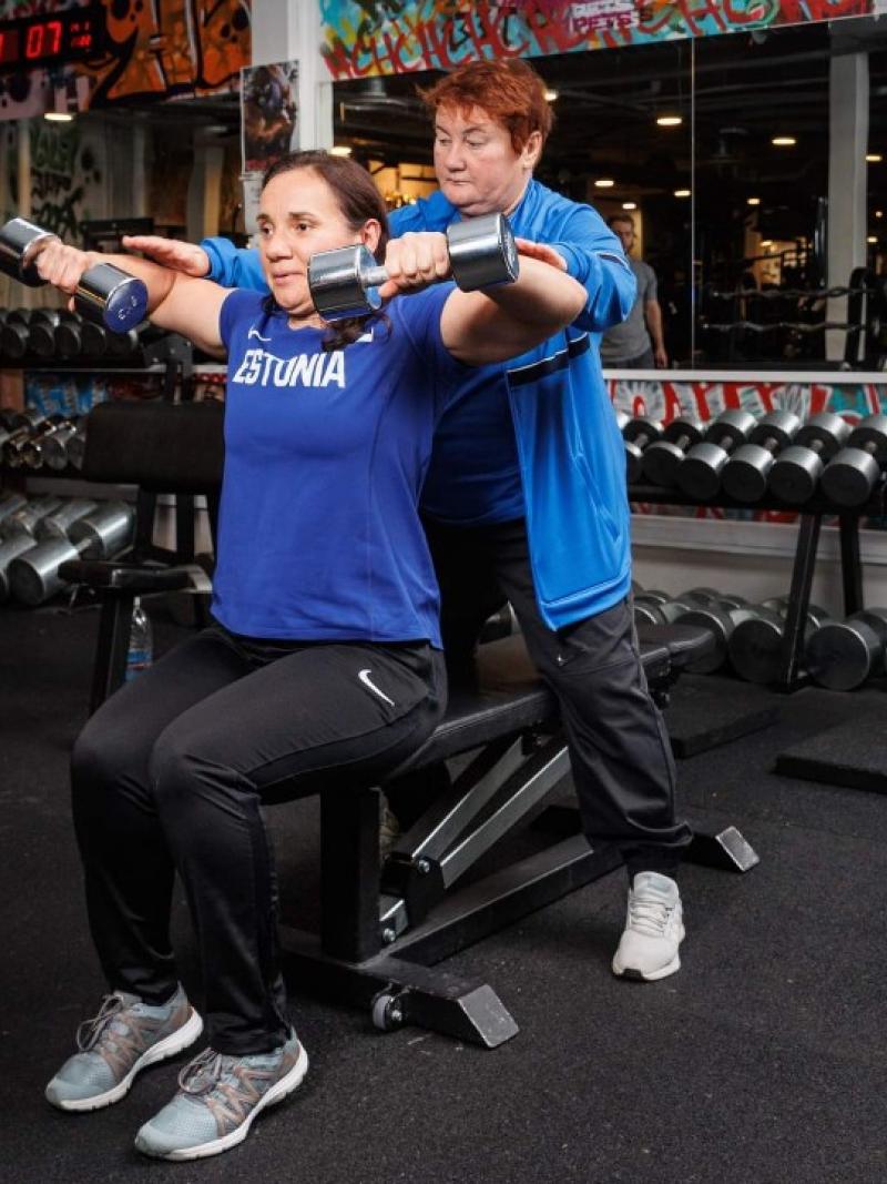 A female athlete in an Estonian T-shirt lifts two dumbbells in a gym as an older woman spots her from behind.