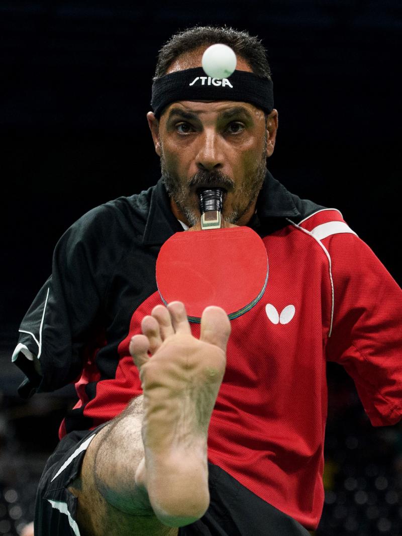 A male Para table tennis player holds a racquet with his mouth and lifts the ball with his right foot.