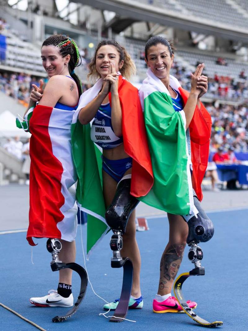 Three female athletes with prosthetic legs posing for a picture with the flag of Italy