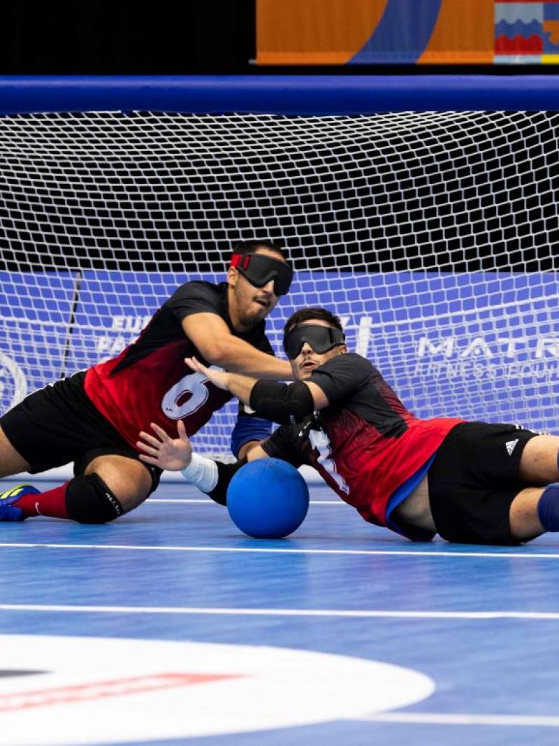 Two male athletes competing in goalball. They are blocking a blue ball in front of the net