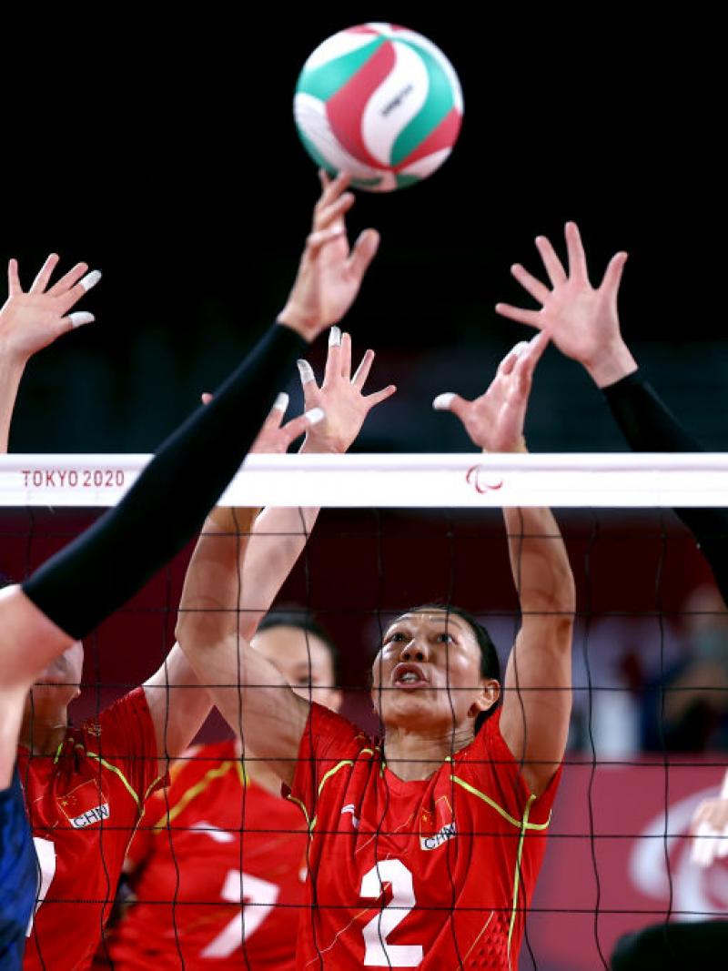 A female athlete tips the ball over the net, while three players try to block it