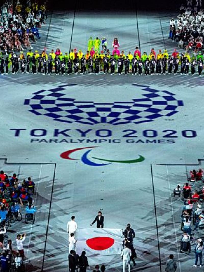 About five people carry the Japanese flag into a stadium during the Closing Ceremony of the Tokyo 2020 Paralympics. The Tokyo 2020 emblem and the Paralympic symbol is depicted on the floor.