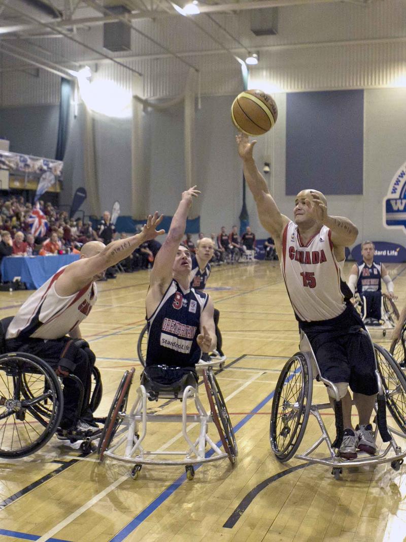 A picture of a man in a wheelchair shooting a Basketball ball between 2 other men in a wheelchair.
