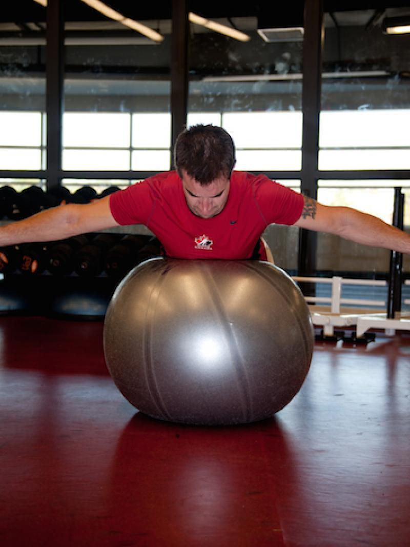 An athlete lies on his stomach on a stability ball with both his arms stretched out to the sides holding weights.
