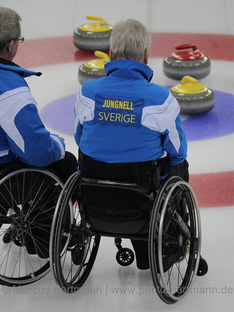 Two men in wheelchair on ice, looking at some curling stones.