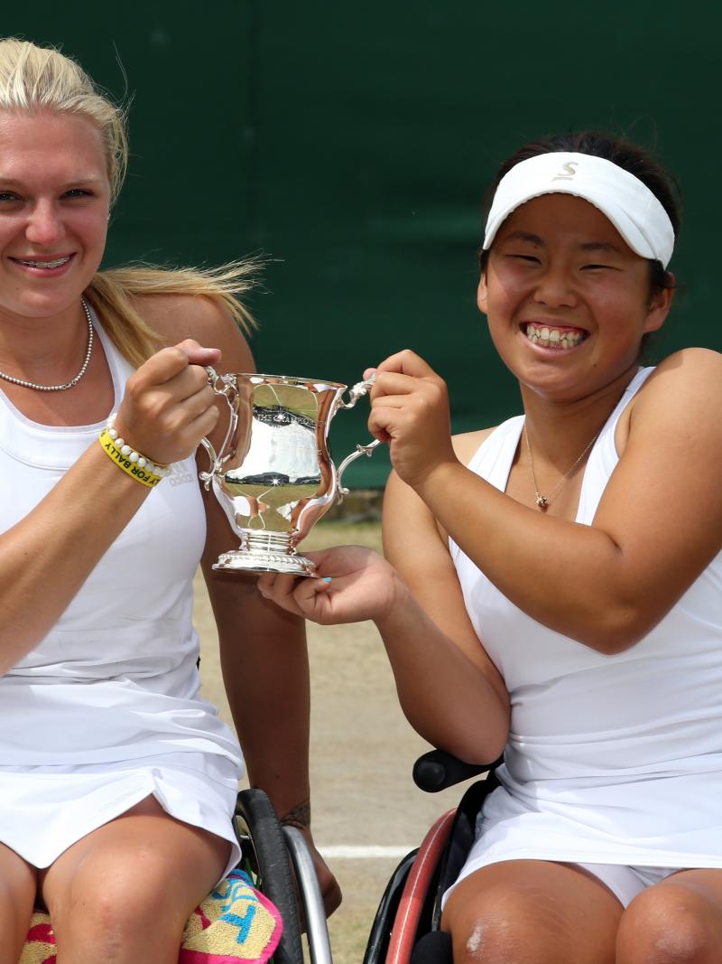 Two women in wheelchairs on a tennis court hold a trophy and smile to the camera.