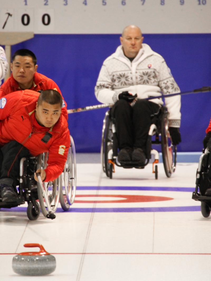 Chinese man in a wheelchair using a stick to throw a curling stone.