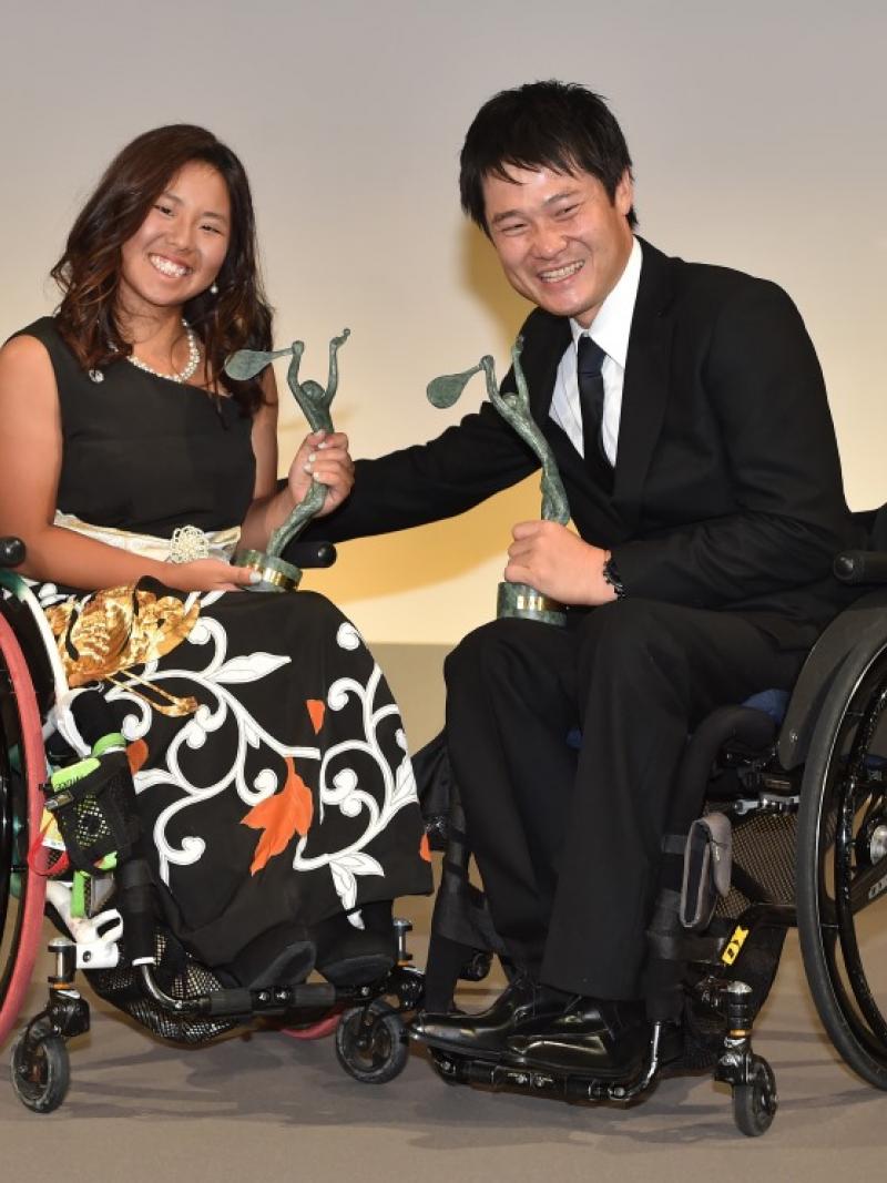 Man and woman in wheechairs holding trophies and smiling to the camera