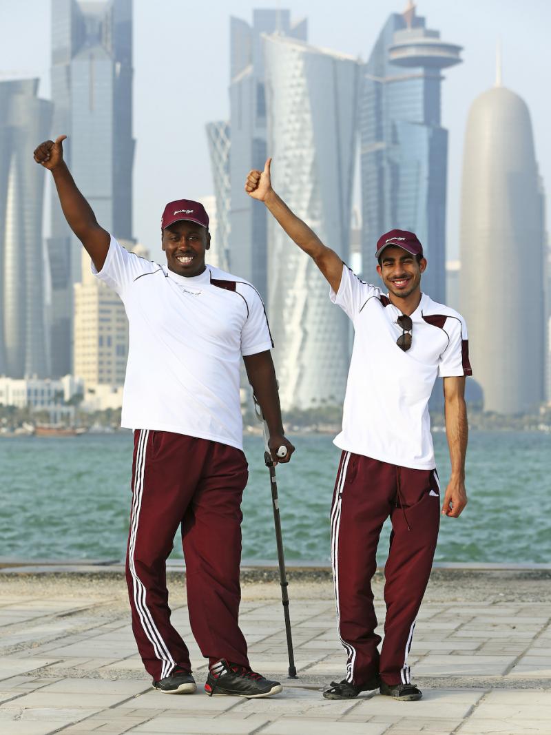 Two men in training suits in front of a skyline, waving