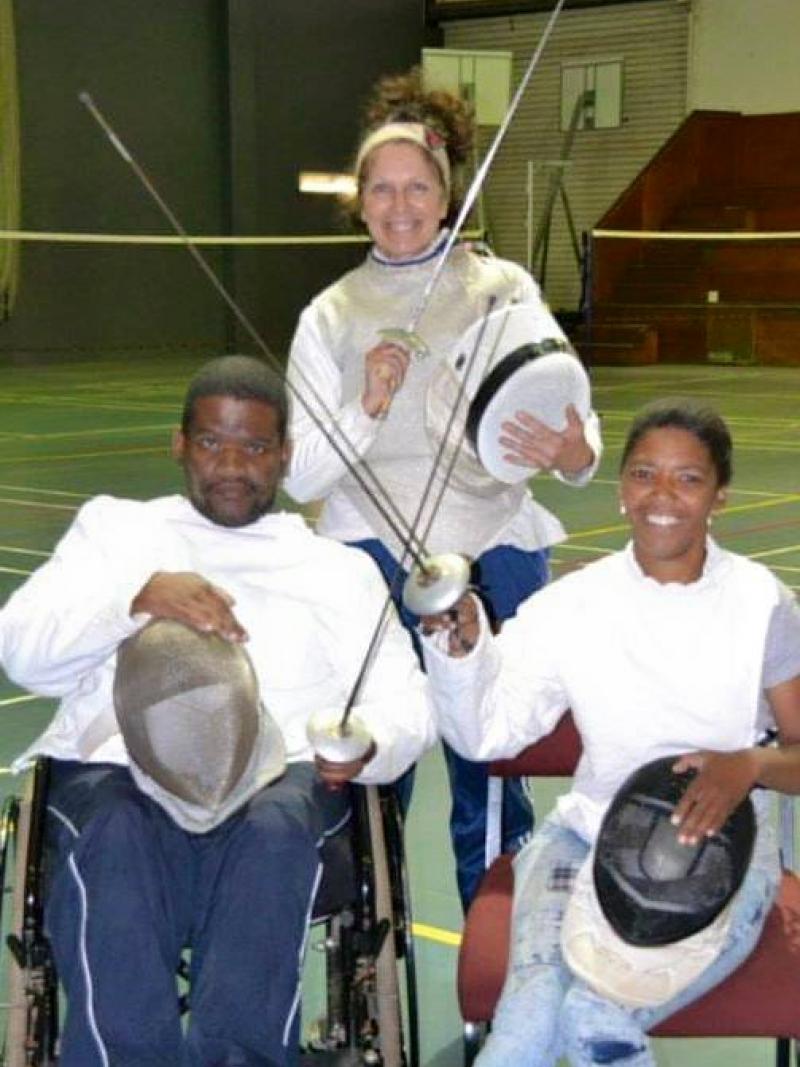 Two fencers in a wheelchair holding a sword and an instructor standing in the background. 