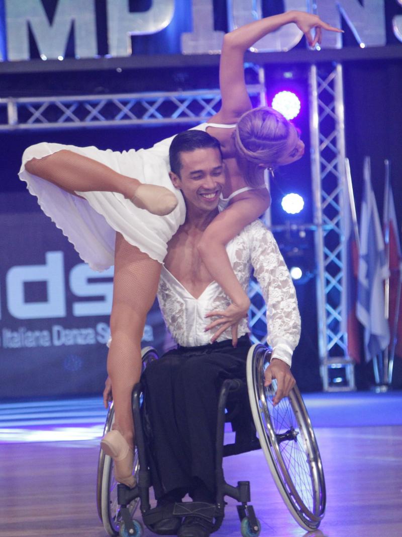 Man in wheelchair holds woman in air while spinning wheelchair.