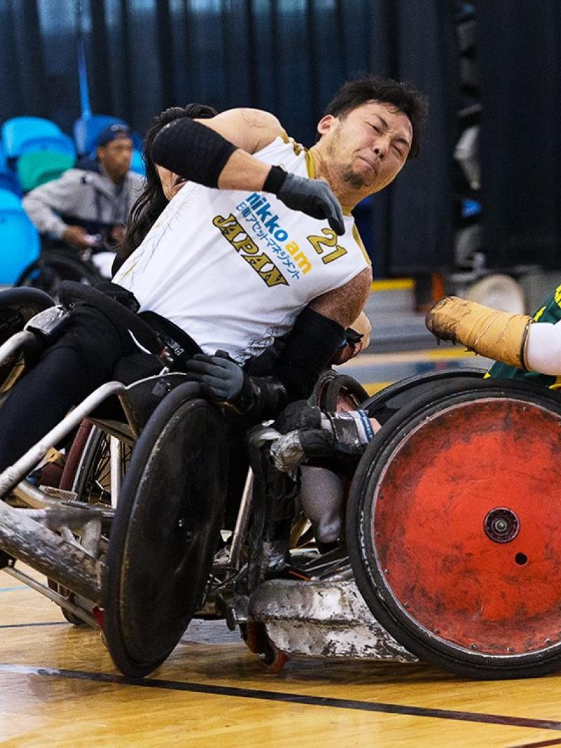 Two men playing wheelchair rugby collide their chairs. 