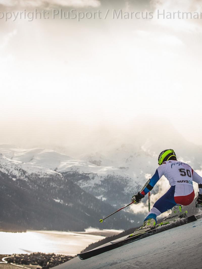 A standing alpine skiier goes down a slope with mountainous backdrop