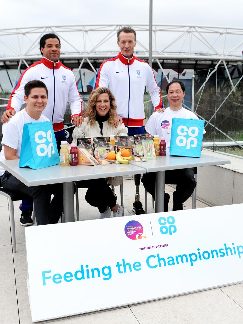 Two men in tracksuits stand behind three people sat at a table. In the background is an athletics stadium.