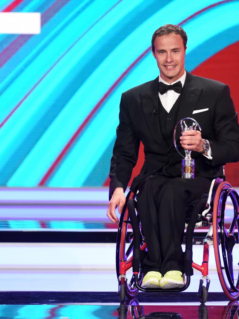 Man in wheelchair dressed in a suit holding a trophy