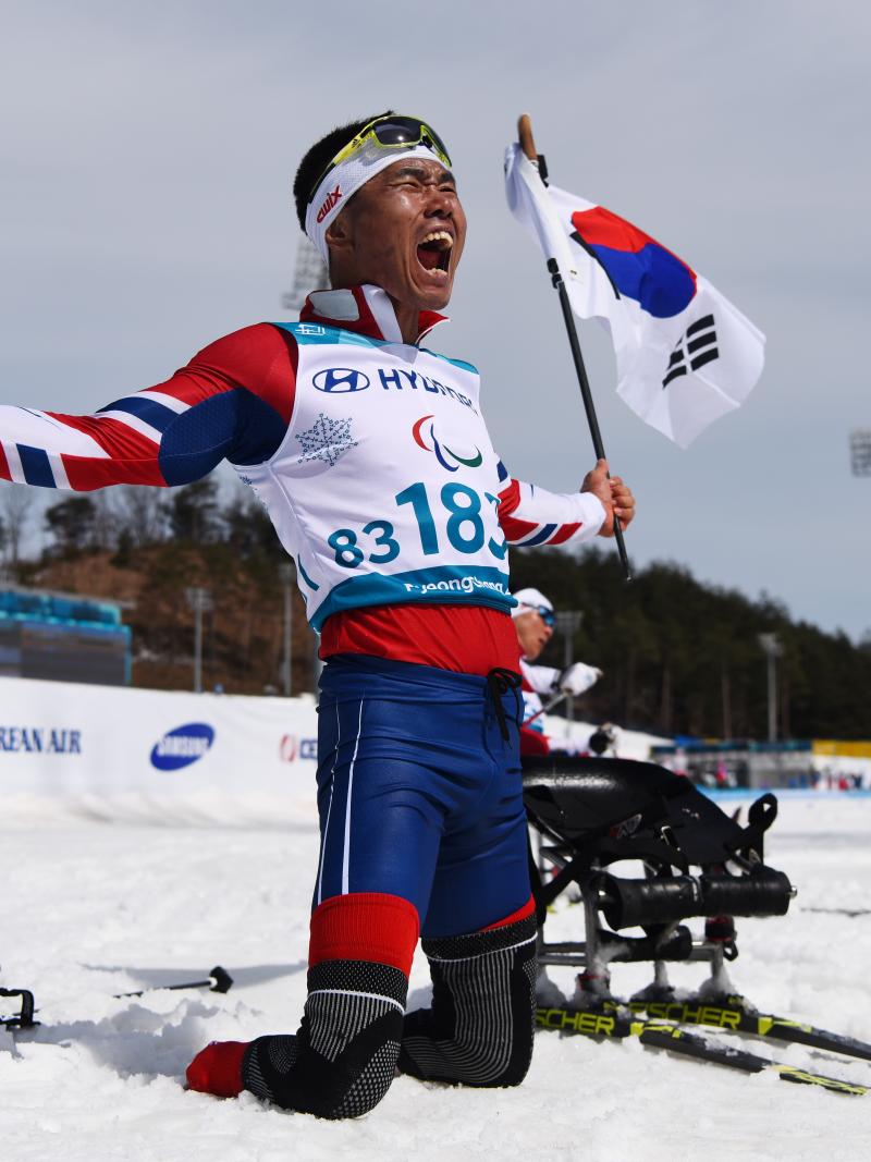 a sit skier celebrates on his knees holding the South Korean flag