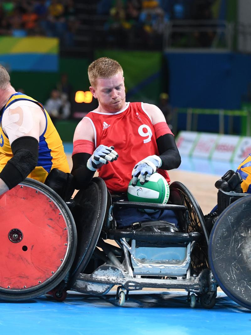 a male wheelchair rugby player holds onto the ball while being challenged by two other players