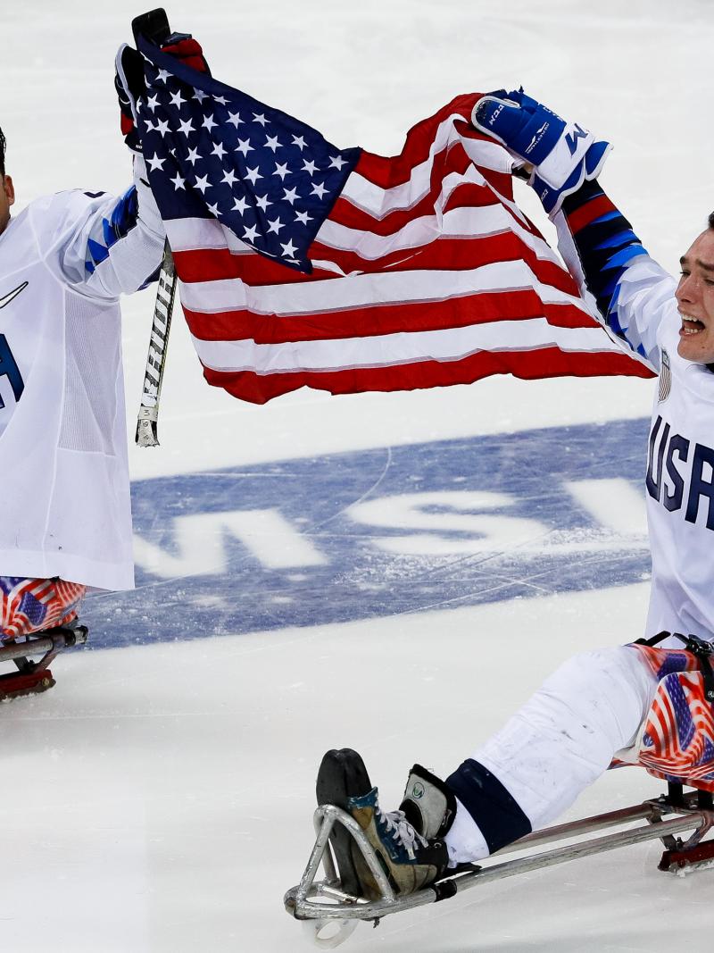 two male US Para ice hockey players on the ice holding a USA flag