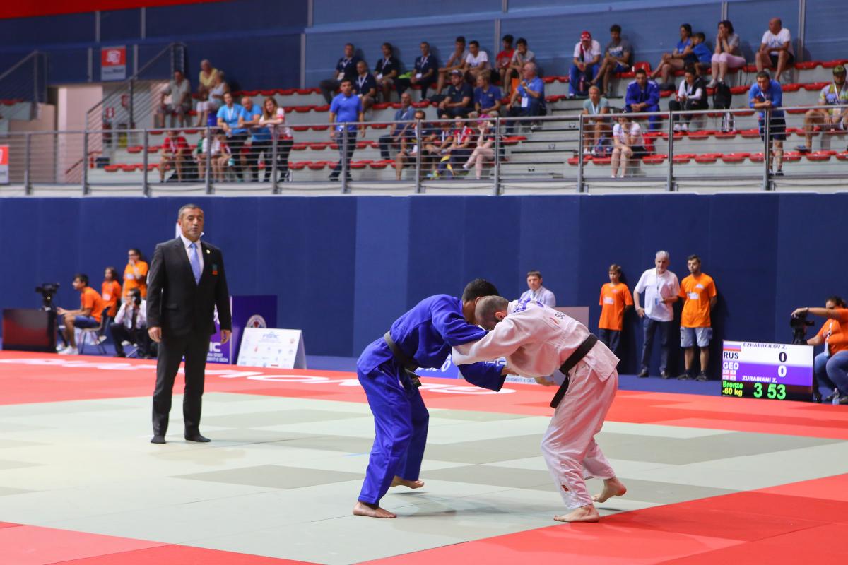 Russian and Georgian judokas grab each other on the tatami as the judge and spectators watch