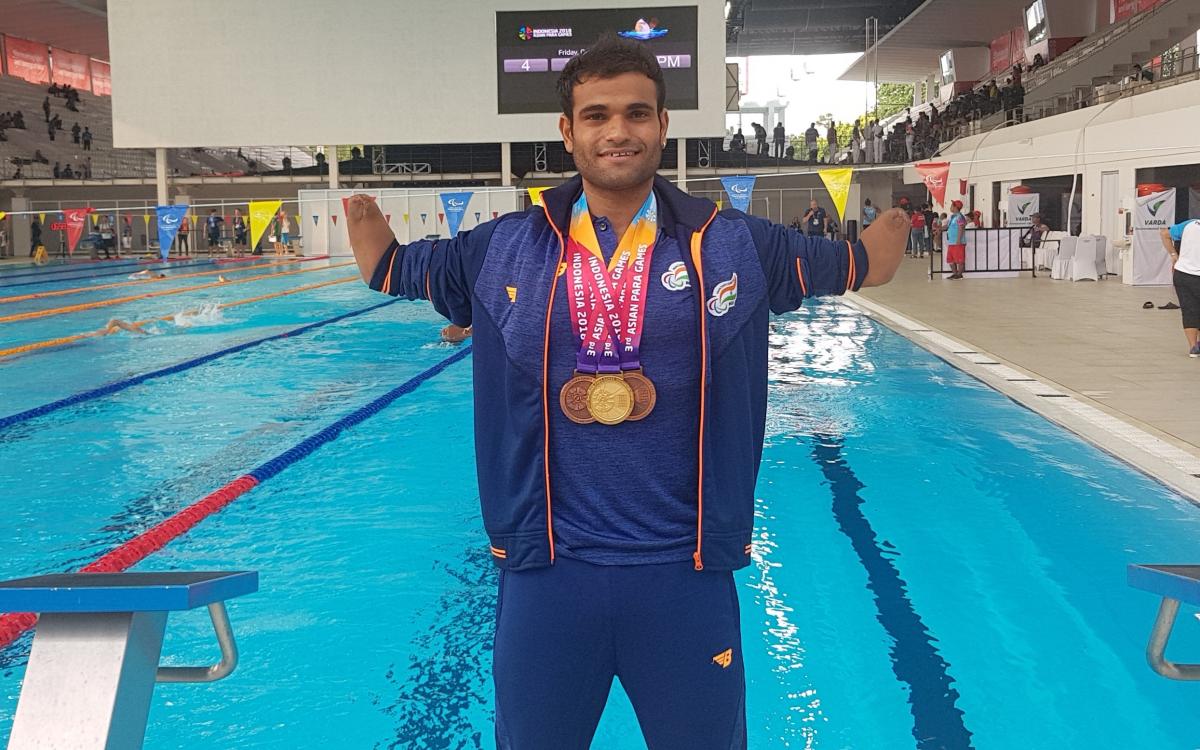 a male Para swimmer standing in front of a swimming pool with medals round his neck