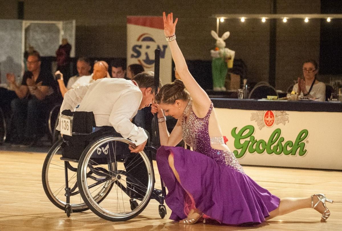 A wheelchair male dancer and a female dancer on a dance floor watched by a group of spectators 