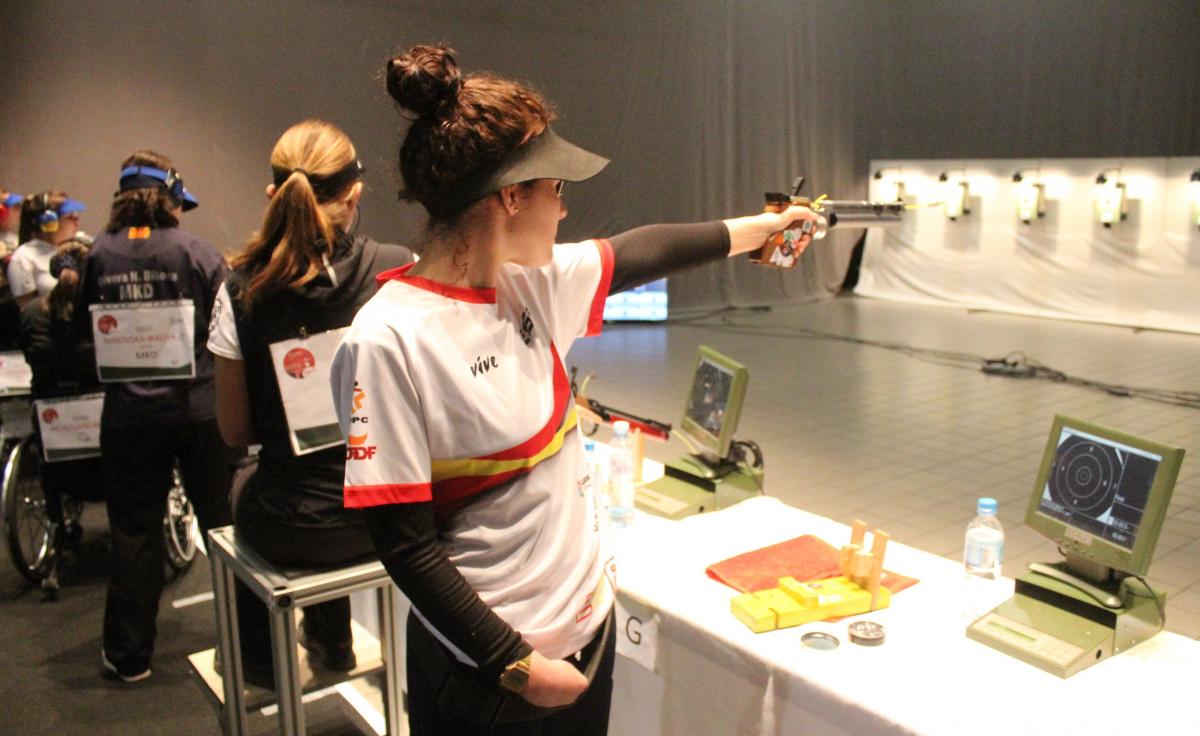 A woman standing and pointing a pistol to a target in an indoor shooting range with four women in the background