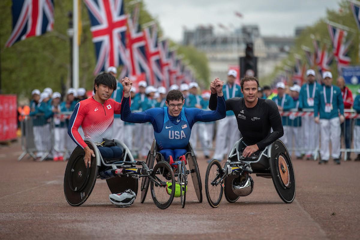 Three men in racing wheelchairs holding hands with the public and flags of the United Kingdom behind them