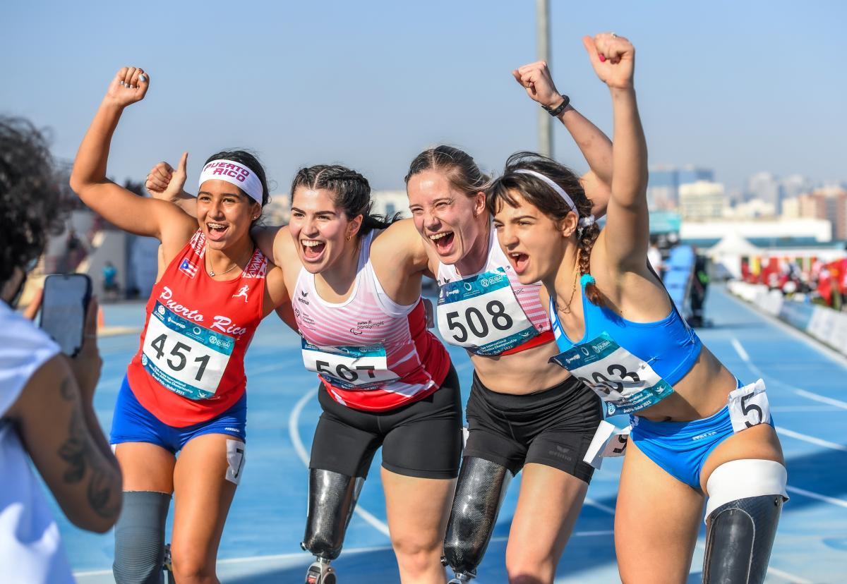 Four women with prosthetic legs hugging each other and smiling to a person taking a picture with a mobile phone