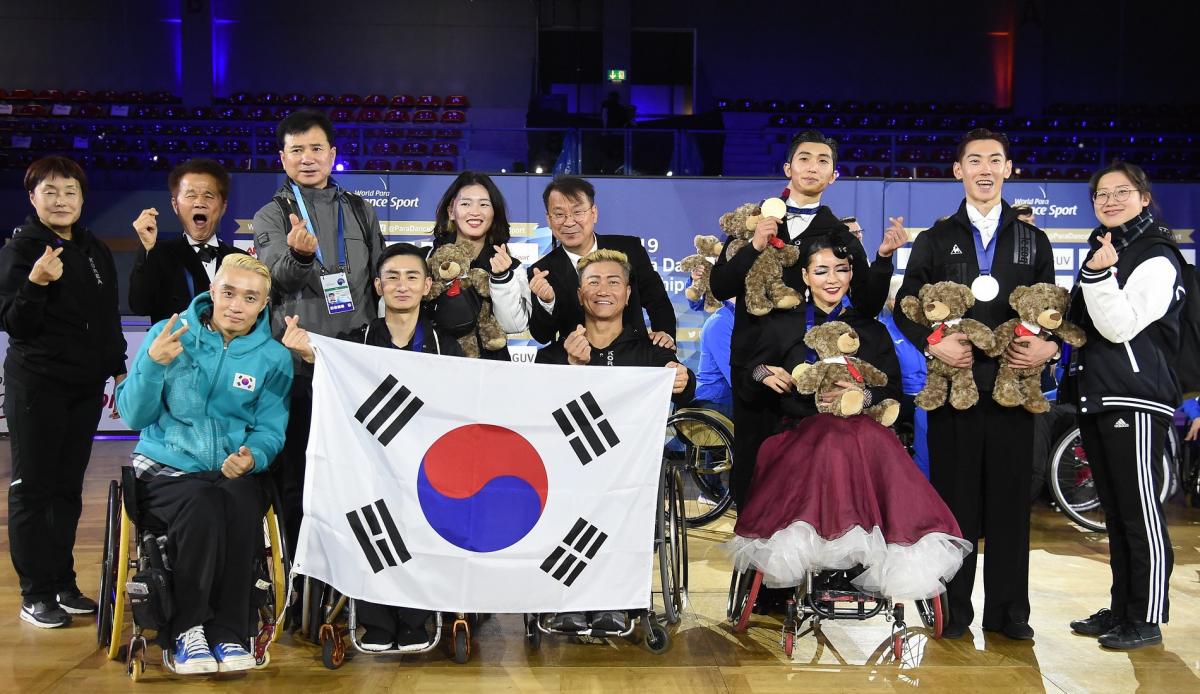 A group of 12 South Koreans, four of them in wheelchairs, posing for a group photo after the Bonn 2019 World Para Dance Sport Championships