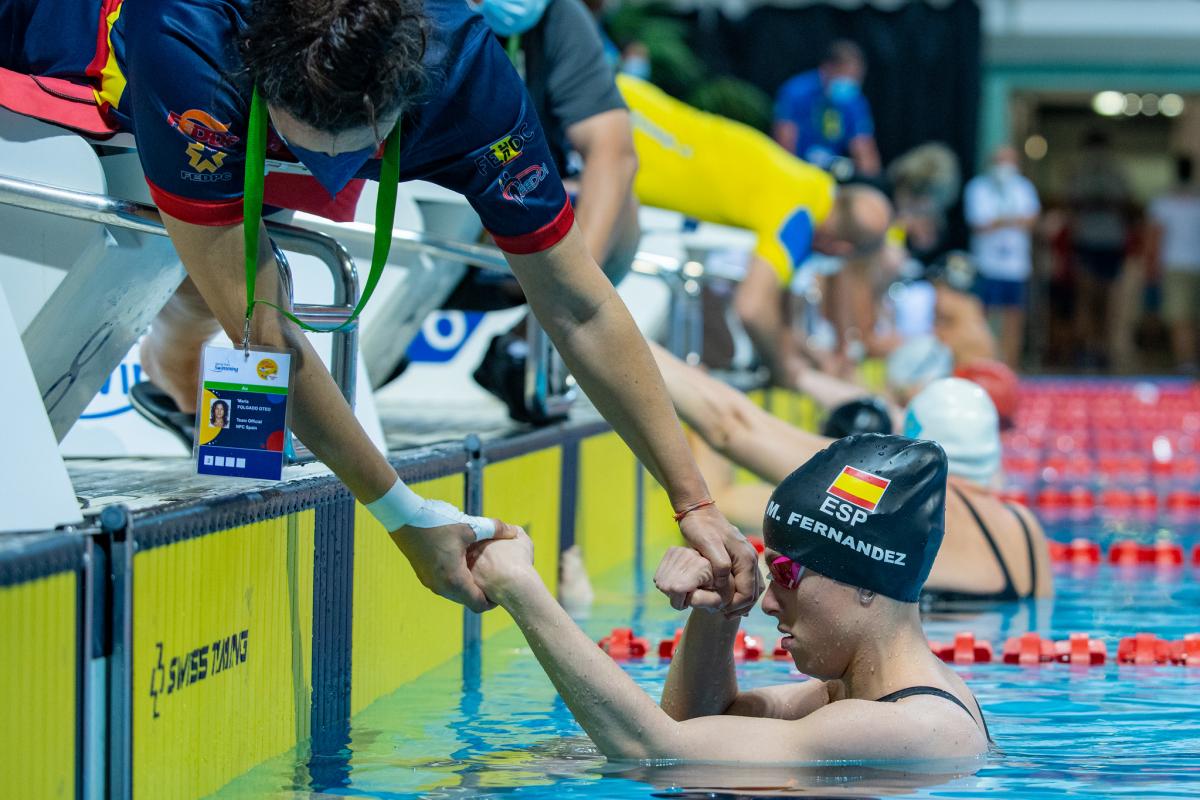 A woman outside a swimming pool grabbing the arms of a female Para swimmer inside the water