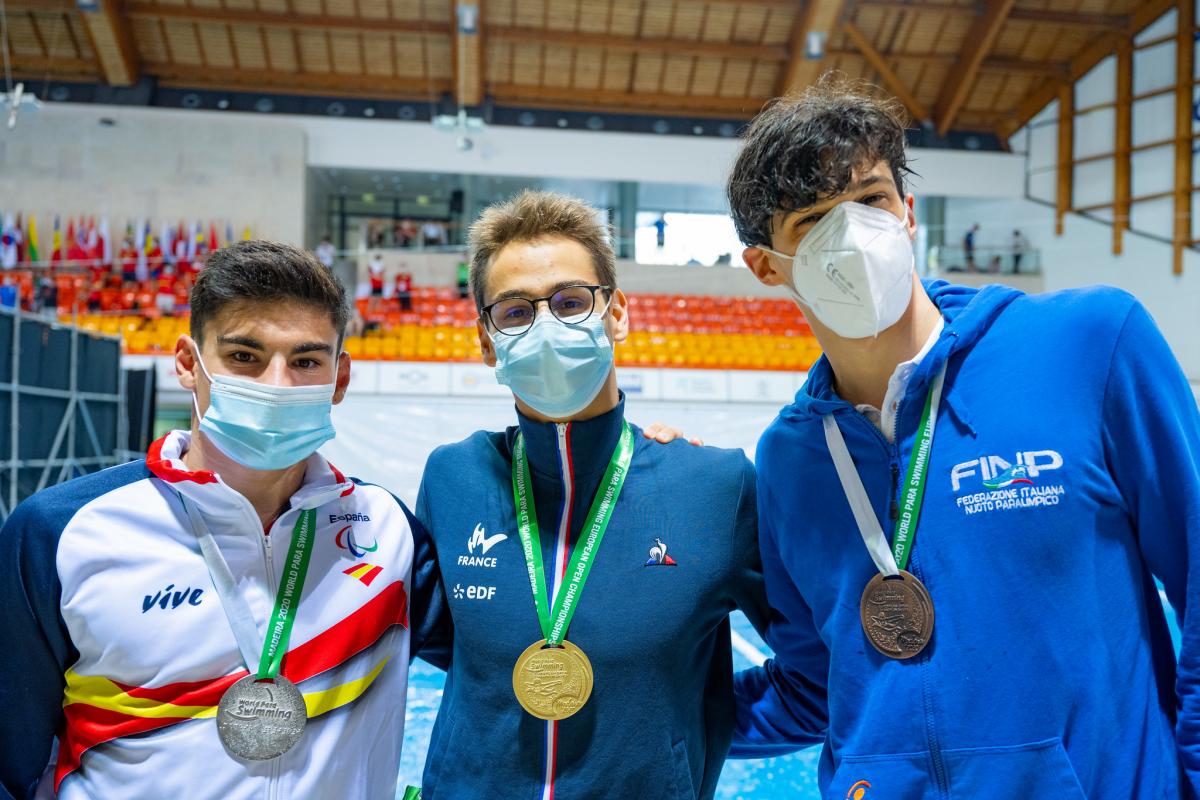 Three men together with their medals in front of a swimming pool