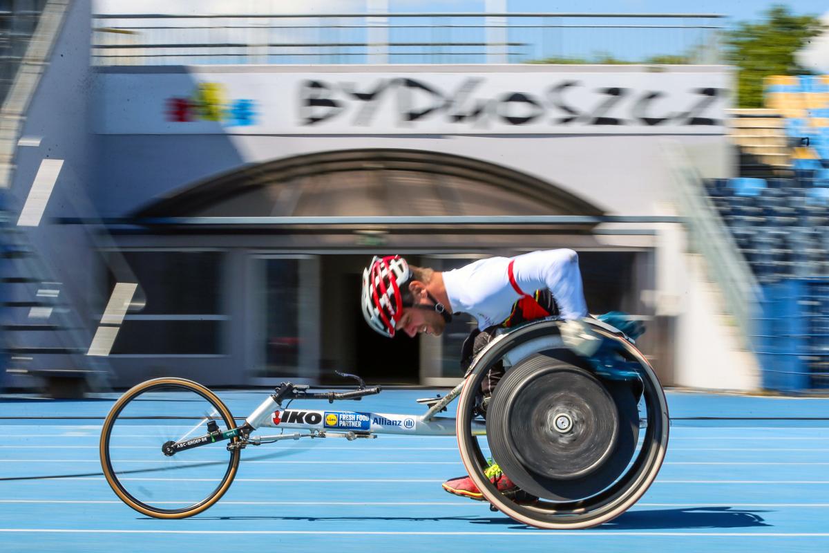 A man in a racing wheelchair on a blue athletics track passing in front of a gate