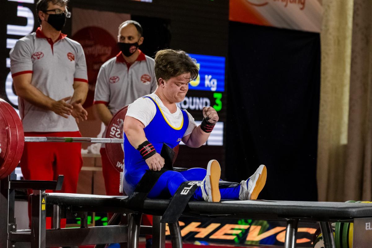 A female powerlifter celebrating seated on the bench press with two men standing behind her