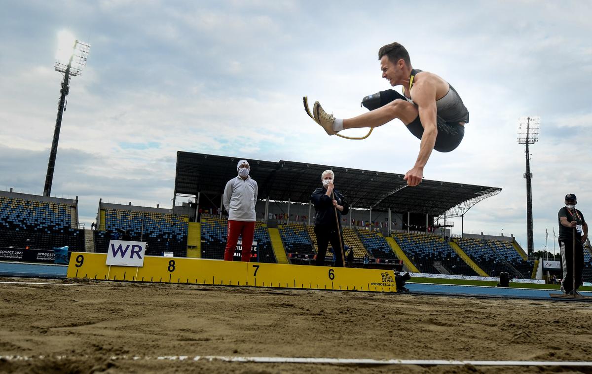 A men with a prosthetic leg jumping in a long jump event observed by three man