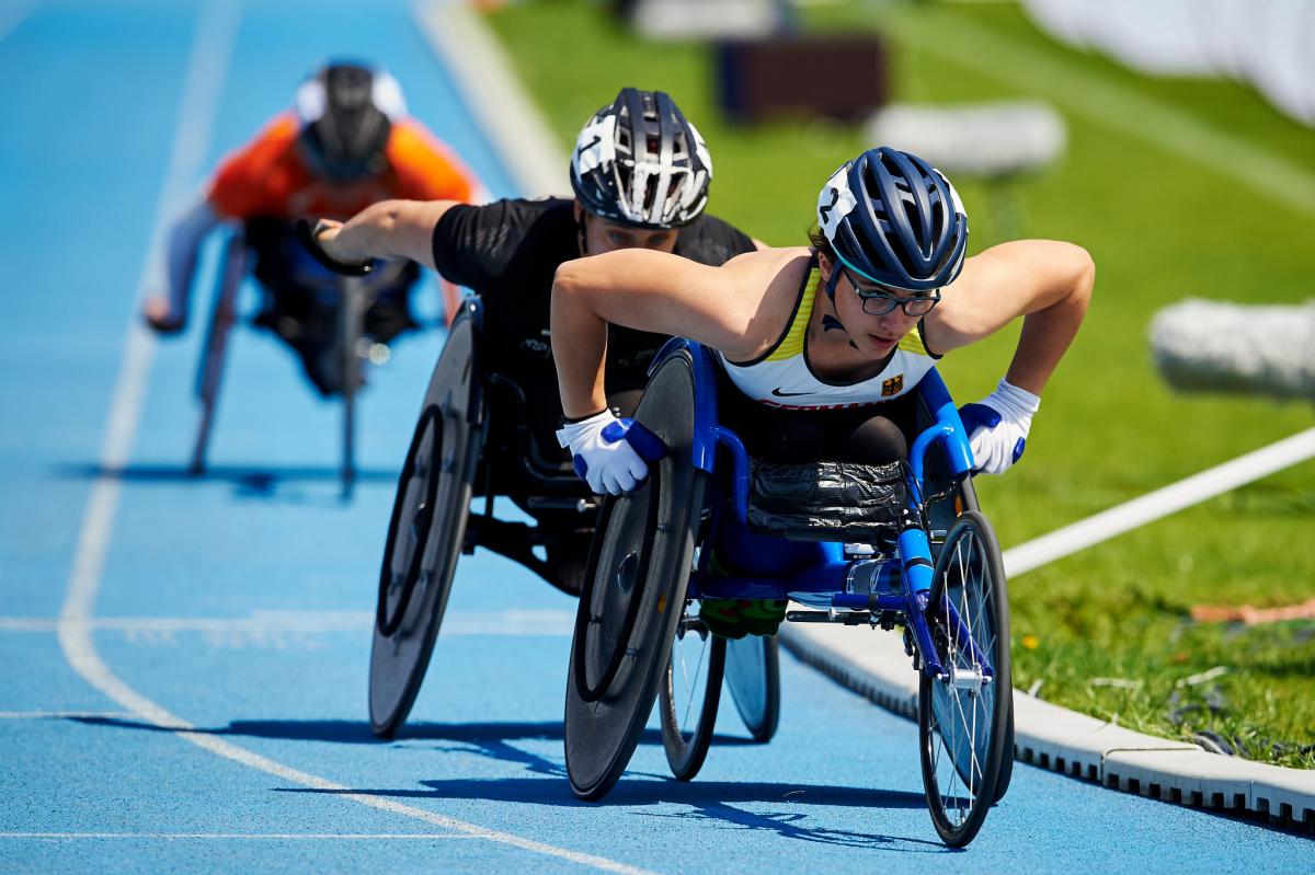 A woman in a racing wheelchair being followed by two athletes during a competition